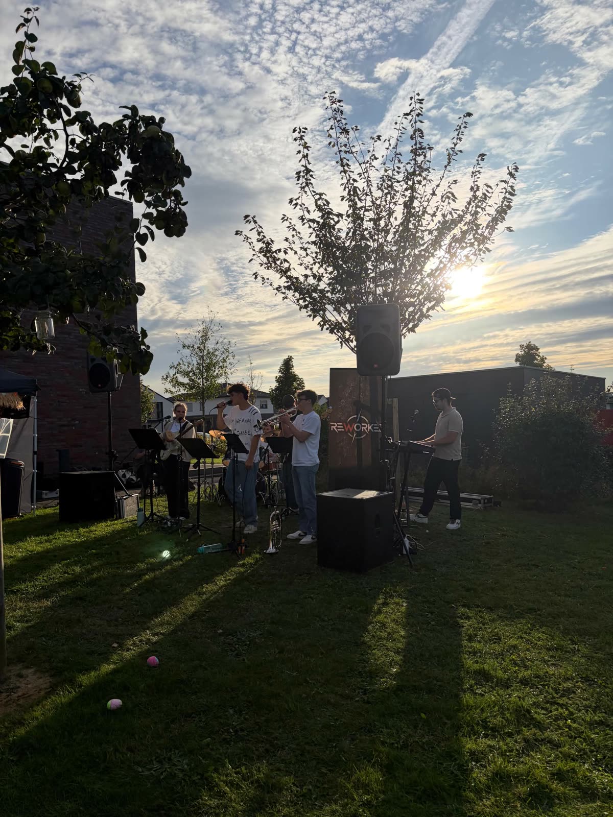 REWORKED beim Keyenberg Sommerfest: die Band spielt in der Abendsonne auf einer Wiese, daneben das REWORKED-Banner und die Boxen